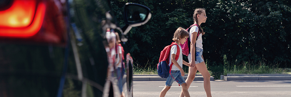 A car idles while two children hold hands and cross the street in a cross walk.